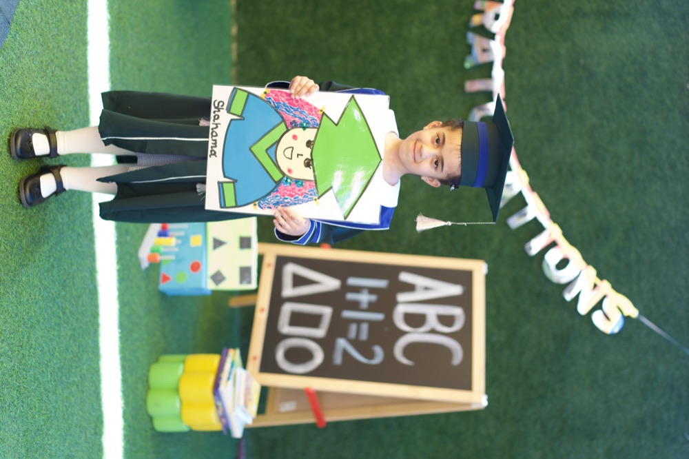 A Little Learners kindergarten graduate in a cap and gown, proudly holding her self-portrait